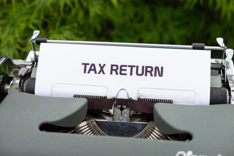 A person sorting tax forms and receipts at a kitchen table, using a calculator and a laptop.